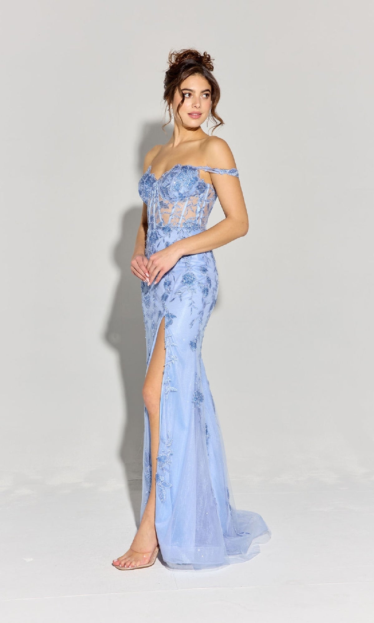 A woman is wearing the Eureka EK181 Off-Shoulder Long Lace Prom Dress in light blue, featuring floral lace embroidery and a high slit, standing against a plain light background.